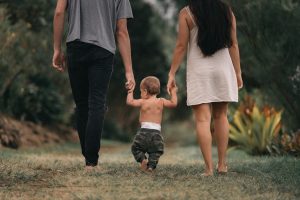Image shows a man and woman holding hands with a toddler between them. All three are walking down a grassy path in their bare feet.