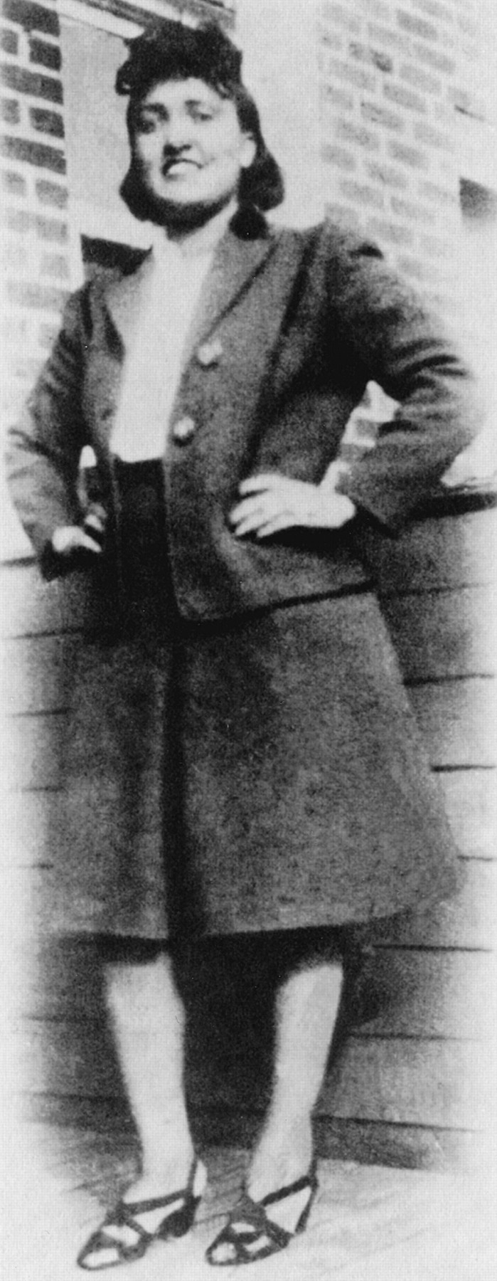 Image shows a black and white photograph of a woman smiling, with her hands on her hips. She is African American, and dressed in the style of the 1940s in a skirt and blazer.