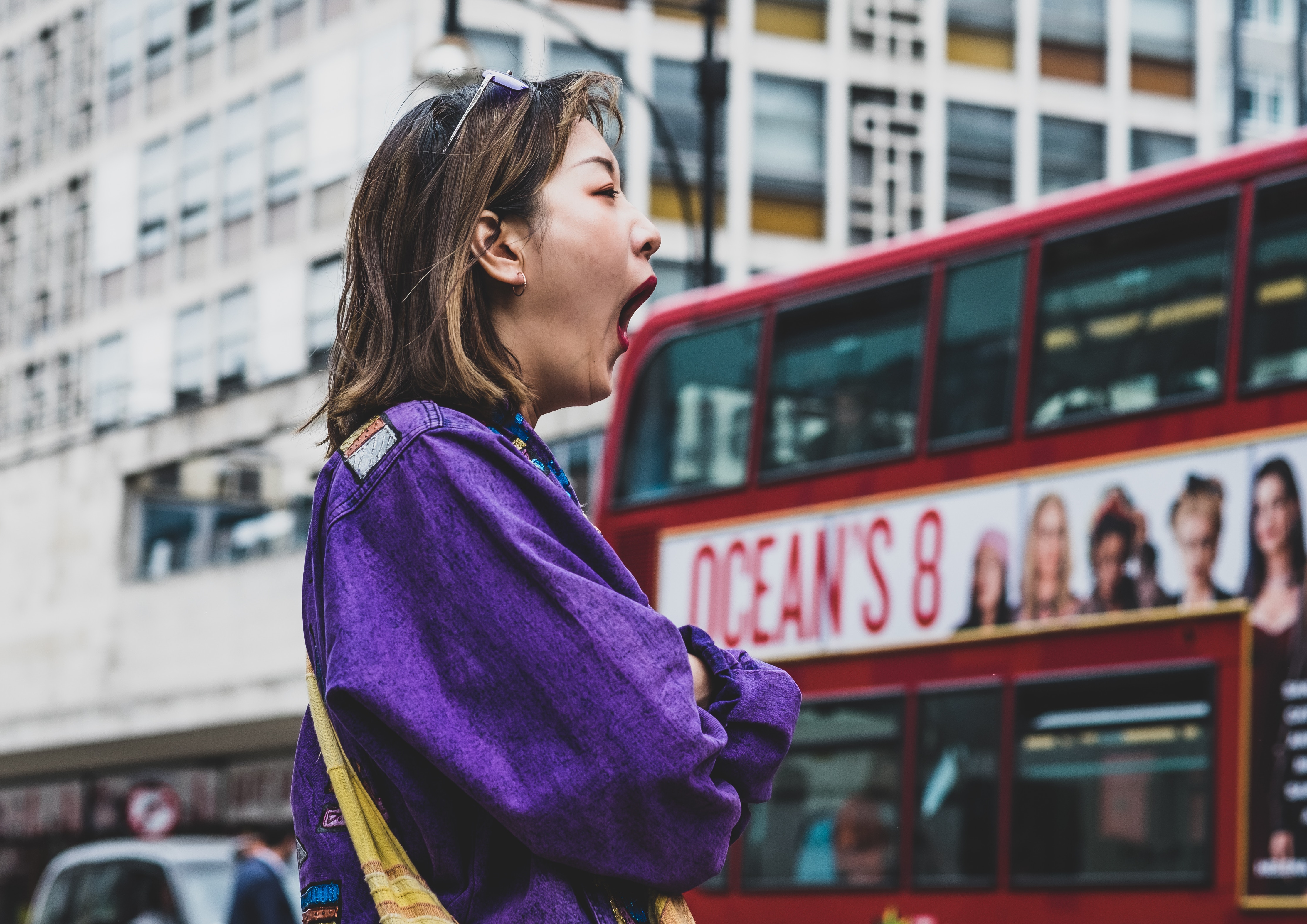 Image shows an Asian woman standing at a bus stop. She is yawning.