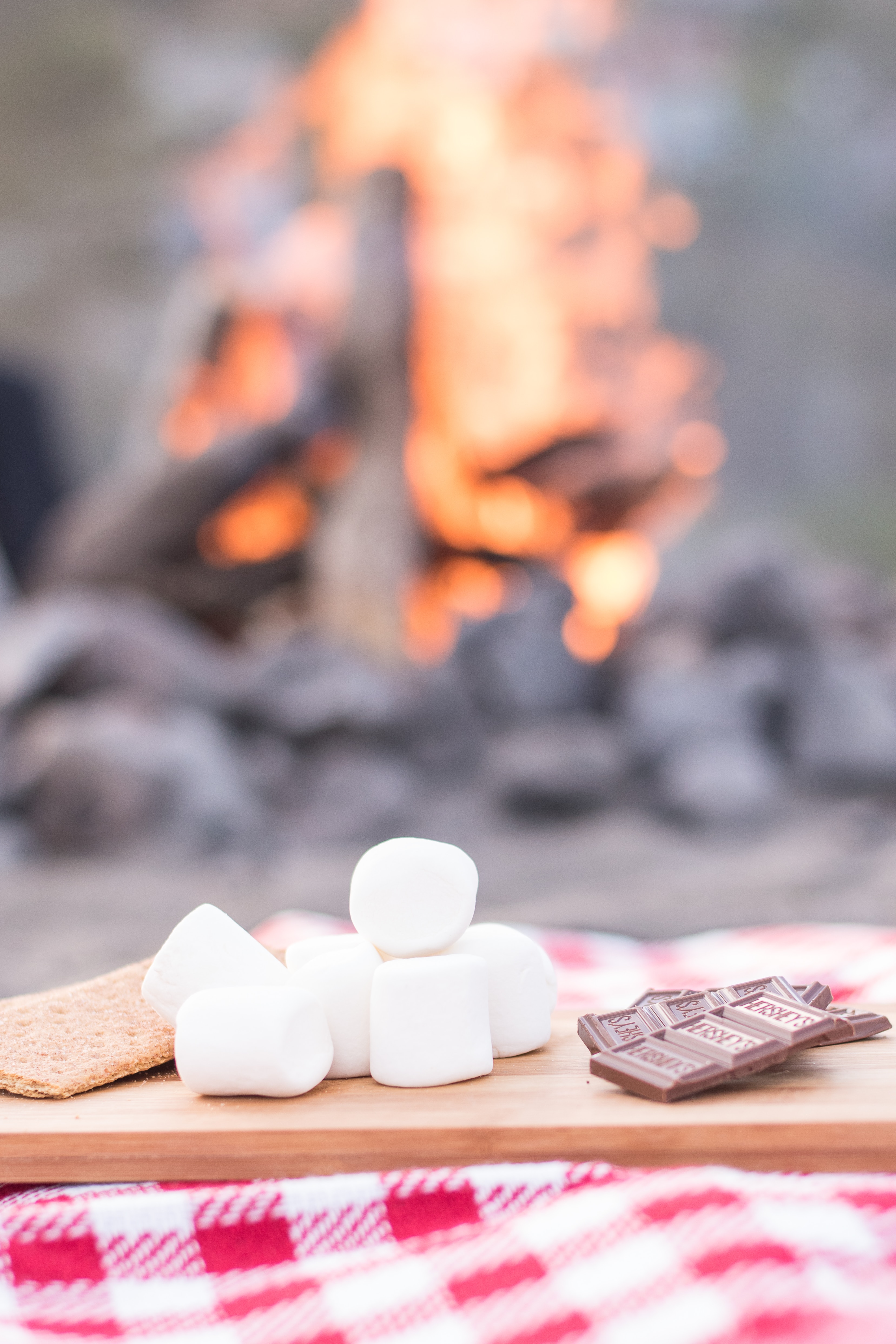 Image shows a photo of the ingredients for smores sitting on a table. In the background, a campfire is burning.